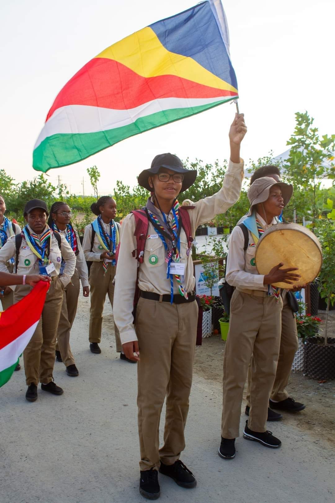 WSJ, Walking to the Opening Ceremony, Partecipants, Seychelles A group of young scouts in uniform marching in a parade, with one scout prominently waving the Seychelles flag while another plays a traditional drum.