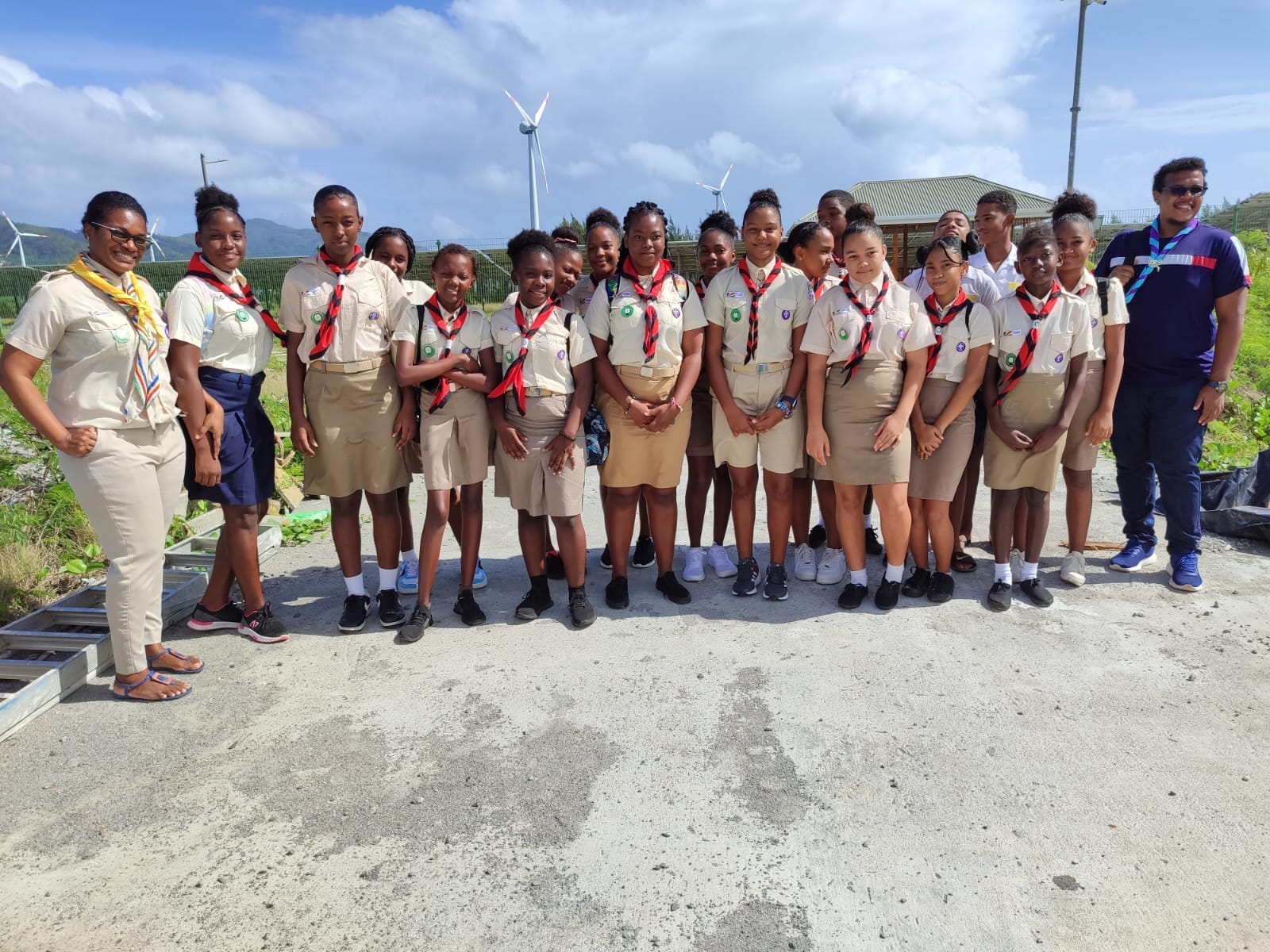 397147954_10233890178424668_7781641390813142865_n Group of young scouts in uniform with leaders, standing outdoors in Seychelles with a wind turbine in the background.