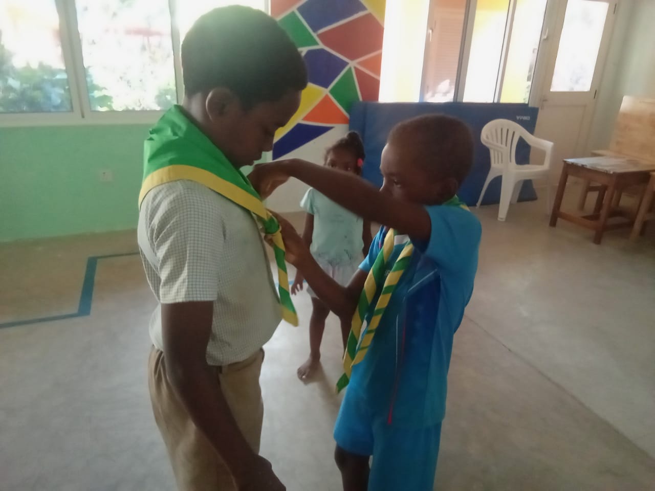 407381769_122127056768062808_4535025070183575548_n A young boy scout helping a boy scout with his neckerchief in a colorful room, preparing for a scout event.