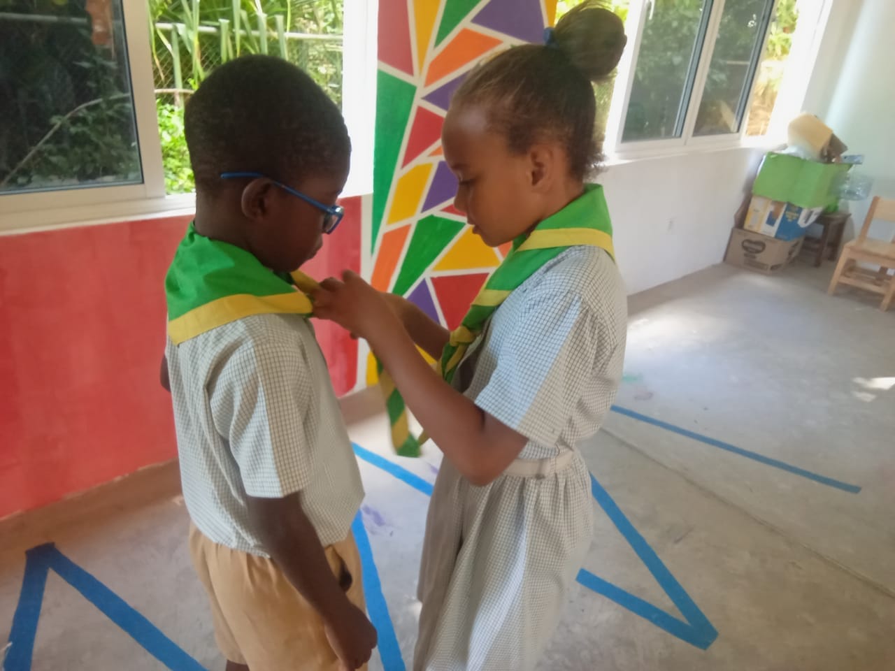 407400701_122127056756062808_296924206076511281_n A young girl scout adjusting the neckerchief of a boy scout in a brightly lit room with colorful wall art, indicative of a scout meeting or activity.