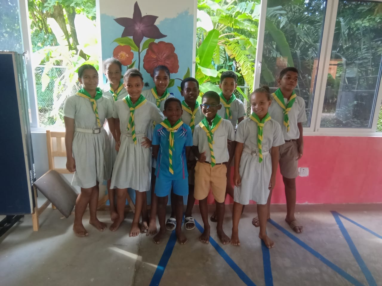 Young scouts in uniform with neckerchiefs standing indoors, in front of a blackboard and a mural, at a scouting event in Seychelles.